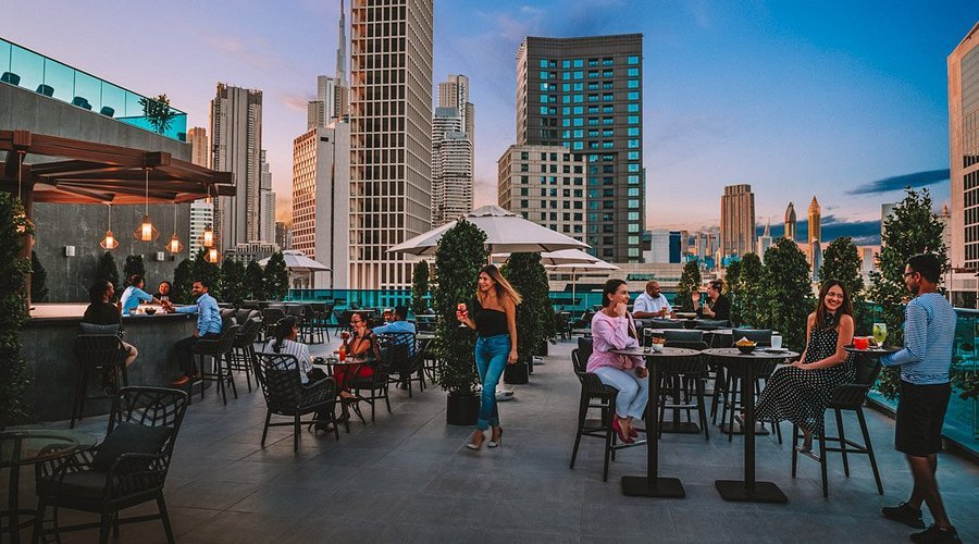 Rooftop bar with people socializing at tables against a city skyline at sunset