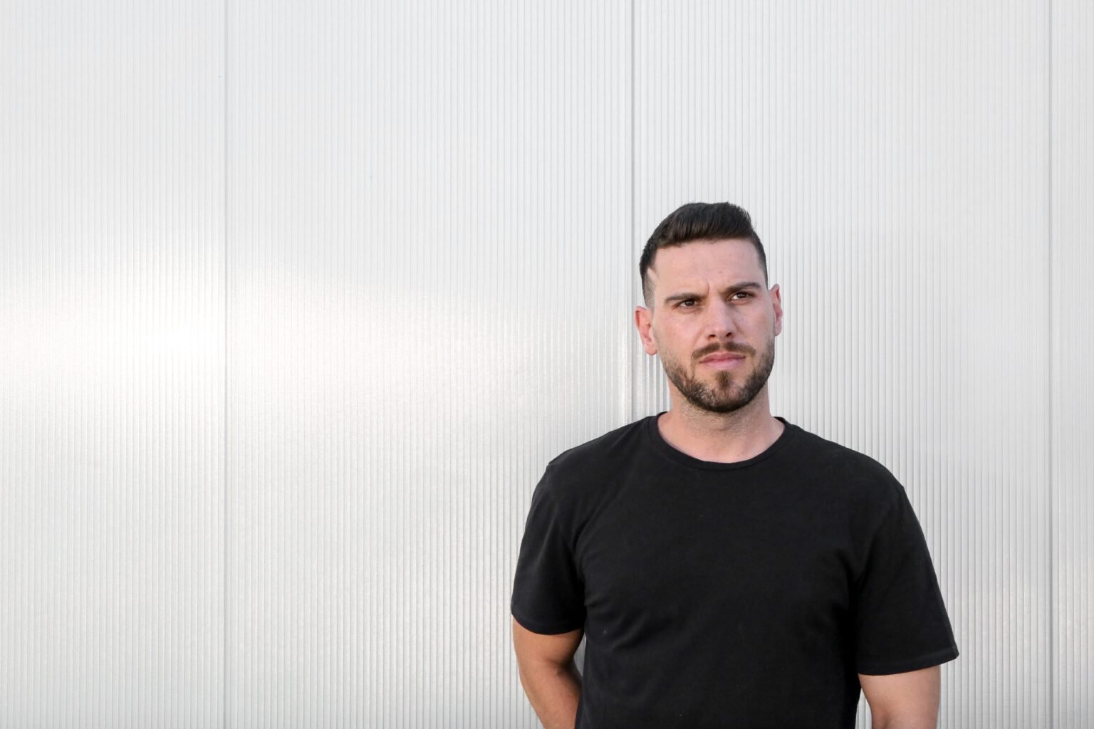Man in a black T-shirt standing against a white ribbed wall, looking off to the side