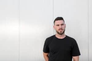 Man in a black T-shirt standing against a white ribbed wall, looking off to the side