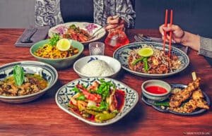 Assorted Asian dishes on a wooden table with two diners reaching in with chopsticks and utensils