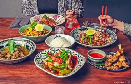 Assorted Asian dishes on a wooden table with two diners reaching in with chopsticks and utensils