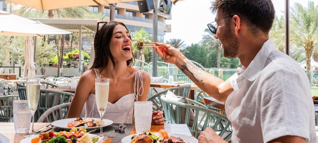 Couple dining outdoors as a man feeds a woman sushi with chopsticks at a sunny patio restaurant