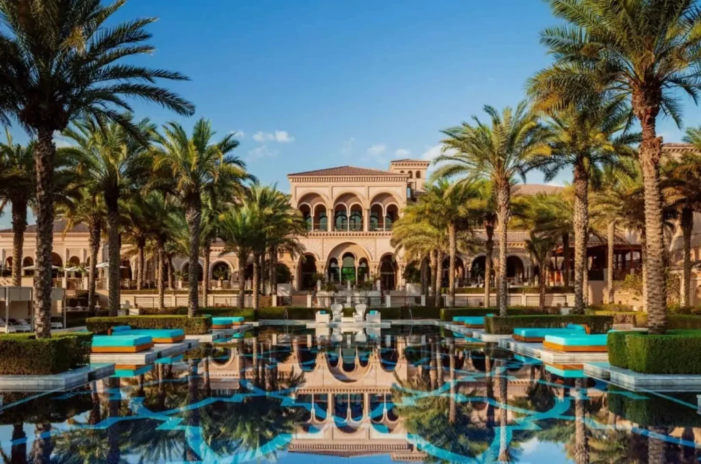 Palm trees framing a resort courtyard with a reflective pool and arched Mediterranean-style building under a blue sky