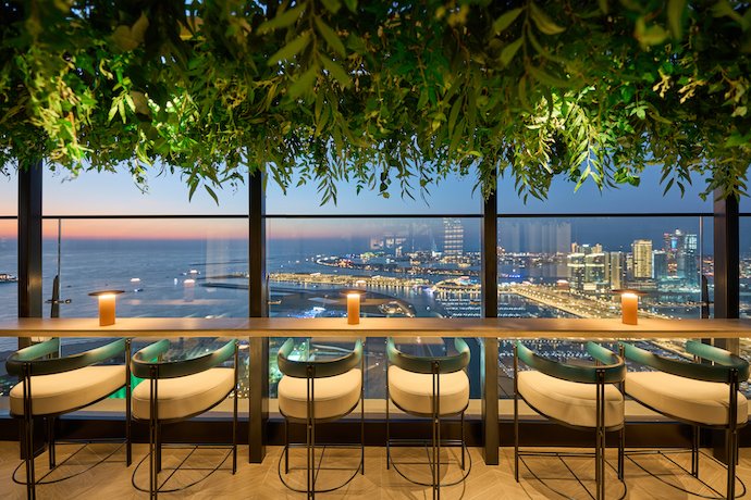 Best Brunch in Dubai: Rooftop bar with stools and hanging greenery overlooking a lit seaside city at dusk