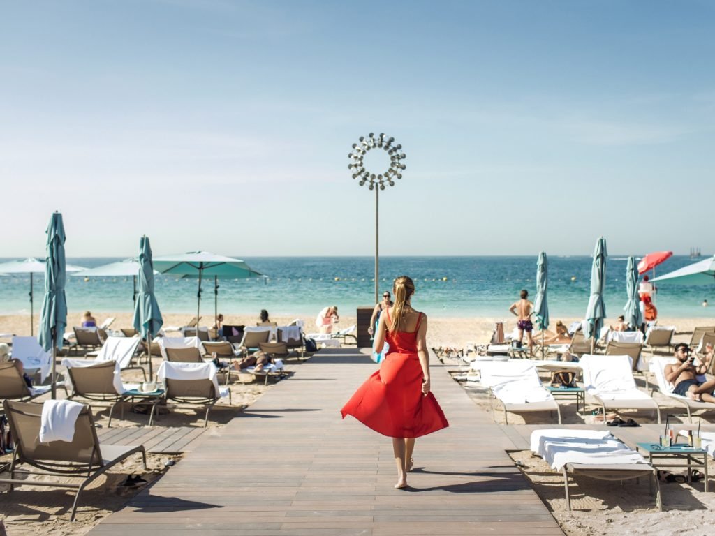 Woman in a red dress walking down a boardwalk toward a sunny beach lined with lounge chairs and umbrellas