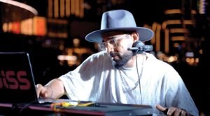 DJ wearing a wide-brim hat and headphones works on a laptop at a nighttime event