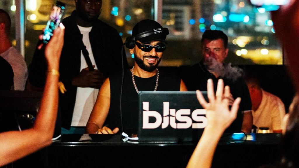 DJ Bliss wearing sunglasses and headphones smiles behind a laptop at a lively nightclub, with raised hands in the crowd