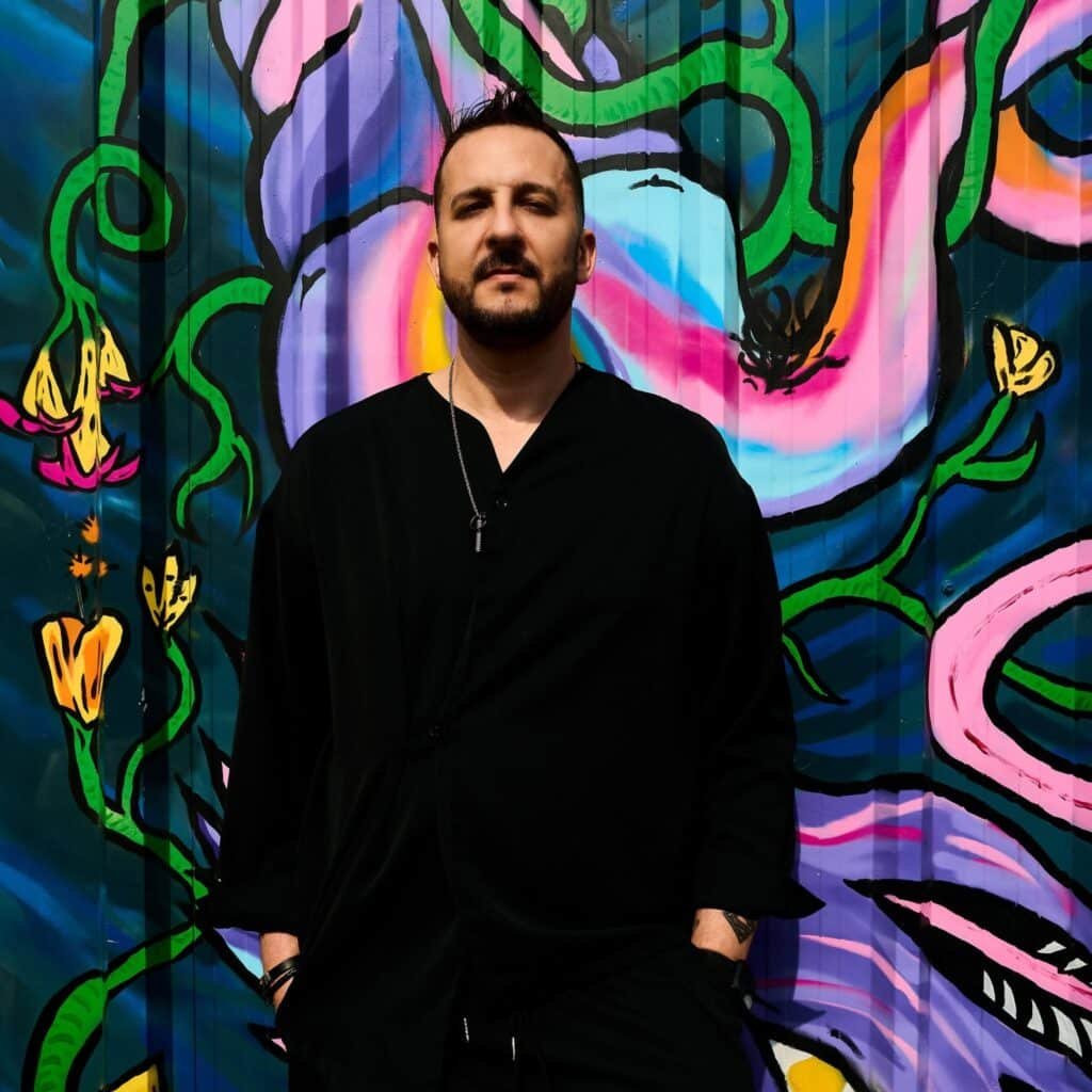 Darko De Jan in black stands in front of a colorful abstract graffiti wall