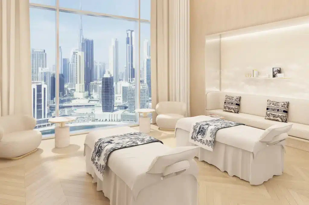 Luxurious beige Dior Haute Wellness spa room with massage beds, lounge chairs, and a city skyline view through a floor-to-ceiling window
