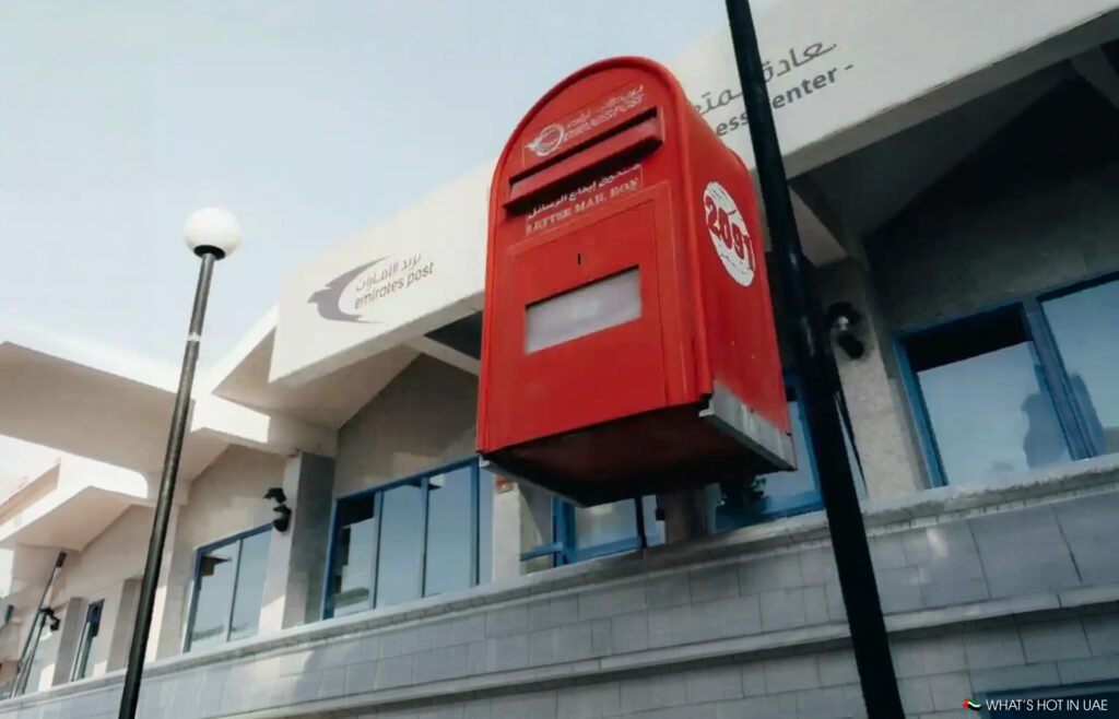Red Emirates Post mailbox mounted outside a building with blue windows and lampposts for when you have no Dubai Postcode