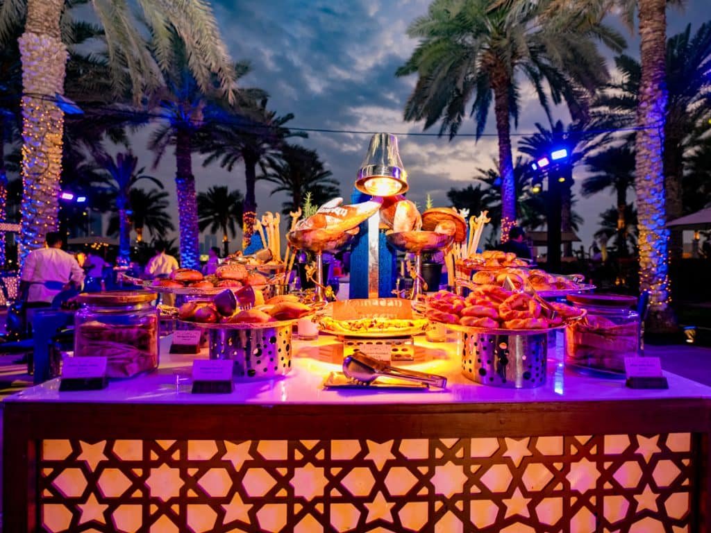 Outdoor seafood buffet under palm trees at dusk, lit in colorful purple and orange lights