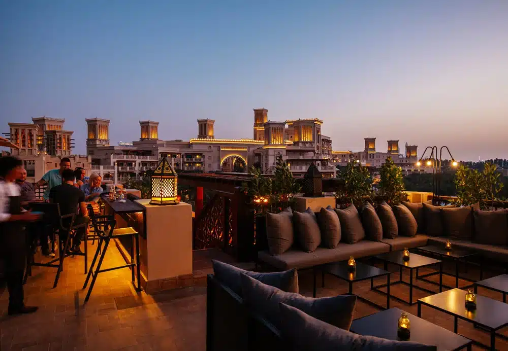 Rooftop lounge at dusk with cushioned seating, lanterns, and guests overlooking lit buildings