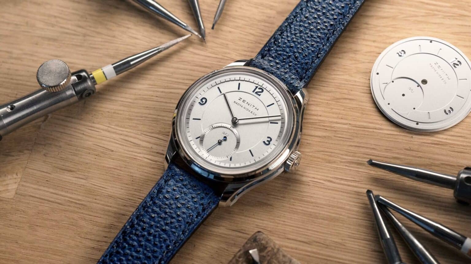Open watch with blue strap on a wooden workbench, surrounded by watchmaker tools and a separate dial