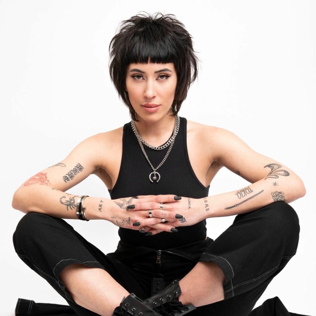 Indira Paganotto with short black hair, tattoos, and black outfit sitting cross-legged against a white background