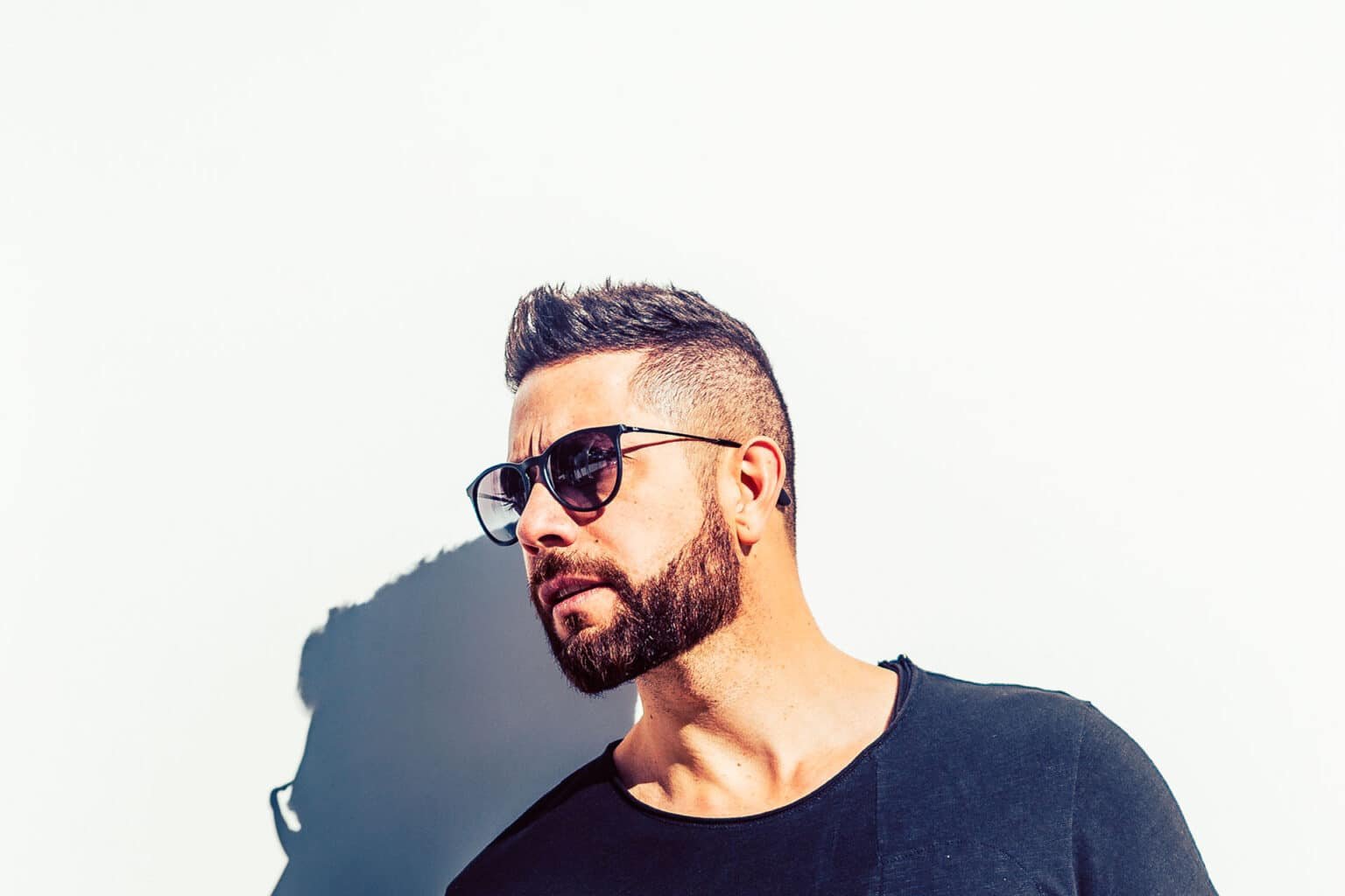 Man with beard and sunglasses in a black shirt against a white wall, with a strong shadow behind him