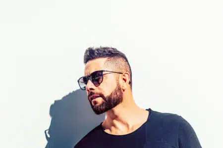 Man with beard and sunglasses in a black shirt against a white wall, with a strong shadow behind him