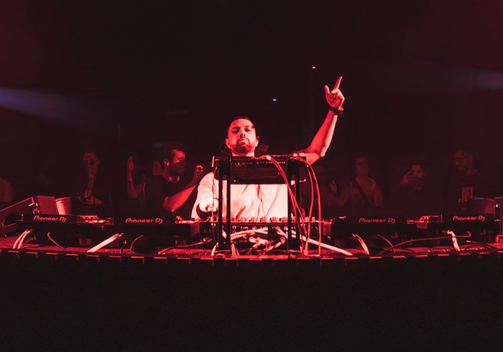 Jean B performing on a dimly lit stage with red lights, raising one hand over a mixer and turntables