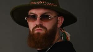 Bearded man wearing a wide-brim hat and tinted sunglasses against a dark background