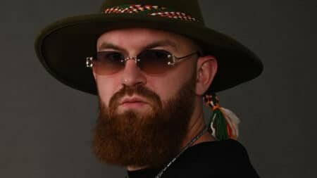 Bearded man wearing a wide-brim hat and tinted sunglasses against a dark background