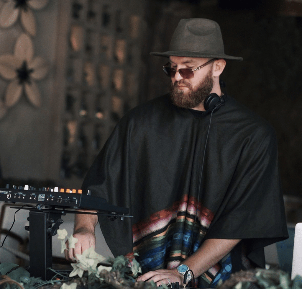 JustBrad wearing a hat and sunglasses, adjusting controls at a mixer in a dimly lit room
