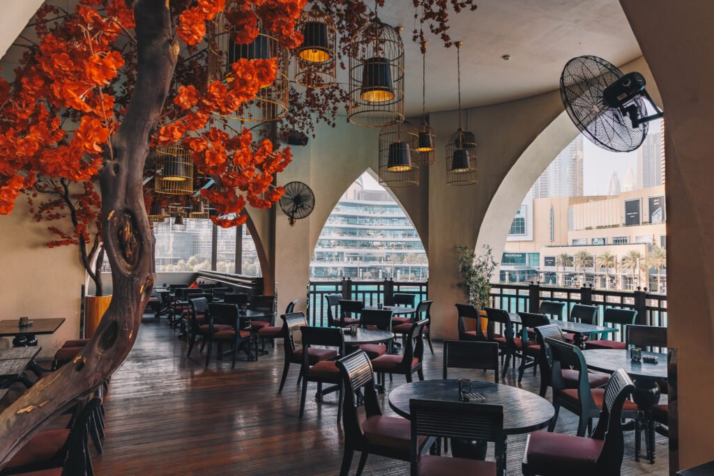 Open-air restaurant terrace with hanging birdcage lights, orange floral decor, and city skyline views