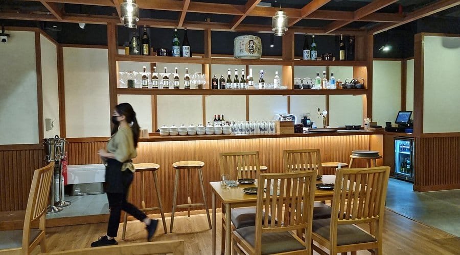 A quiet Japanese-style restaurant interior with wooden tables, stools, shelves of bottles, and a masked server walking by