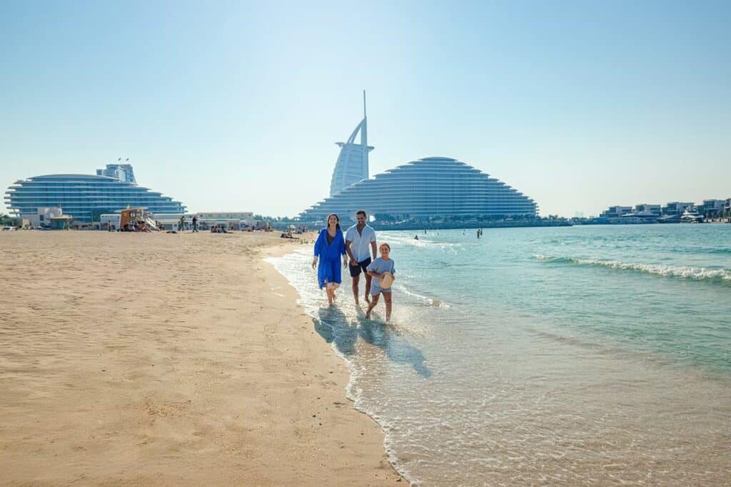 Family walking along a sunny beach with the Burj Al Arab and nearby resort buildings in the background