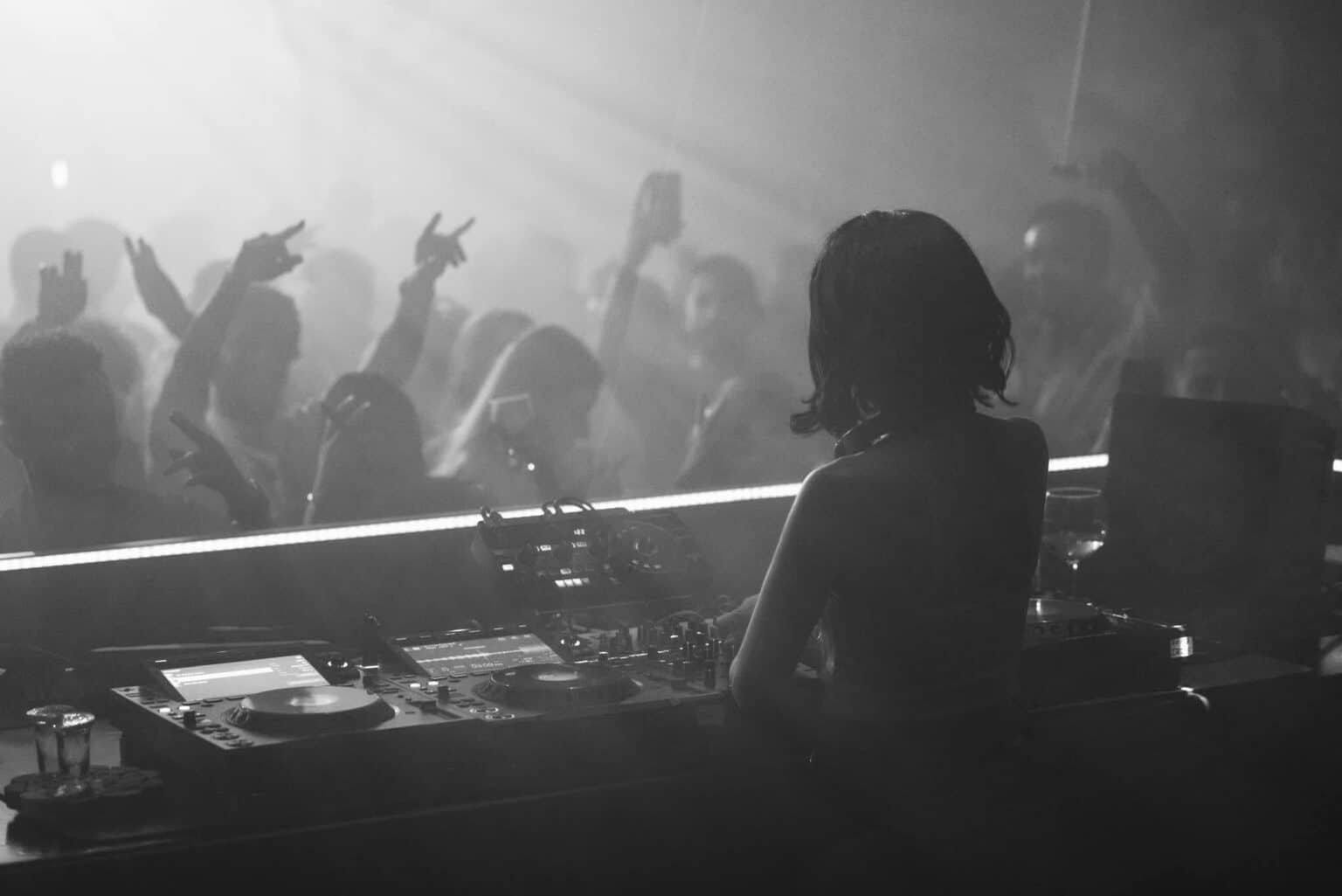DJ performing at a nightclub with a cheering crowd and raised hands in smoky black-and-white lighting