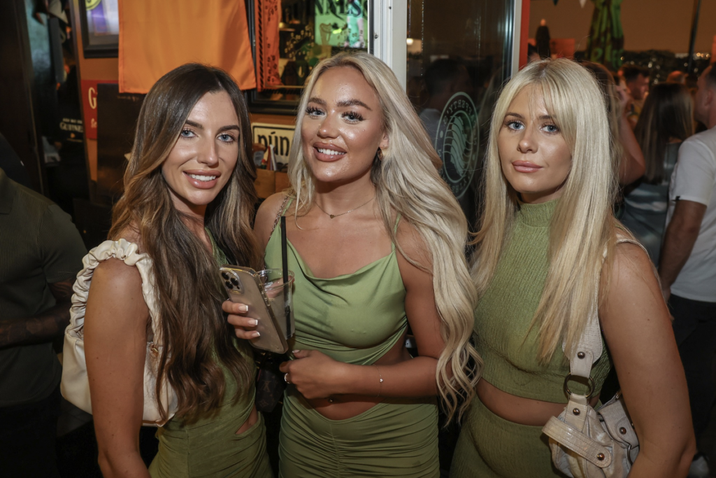 Three women in matching green outfits posing at a crowded nightlife event