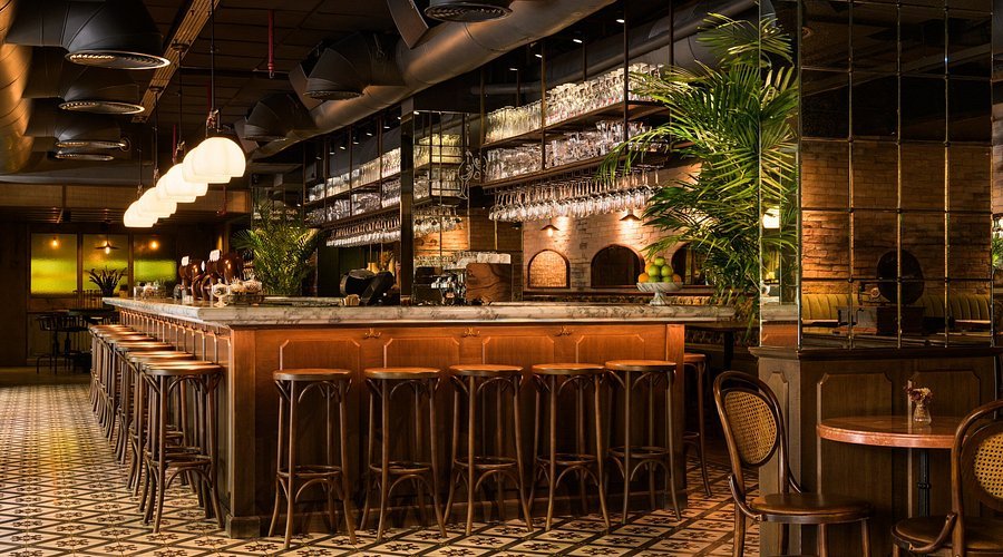 Warmly lit bar interior with a long marble counter, tall stools, hanging glasses, and patterned tile floor