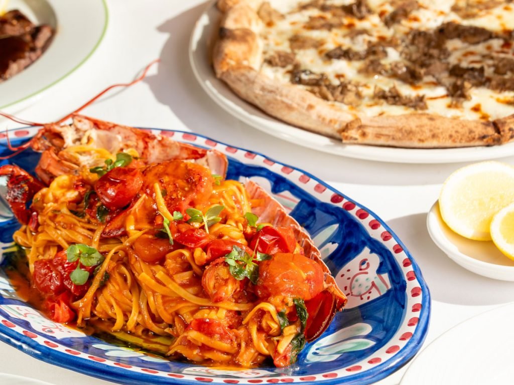 A colorful bowl of spaghetti with tomato sauce and herbs, beside pizza and lemon wedges on a white table