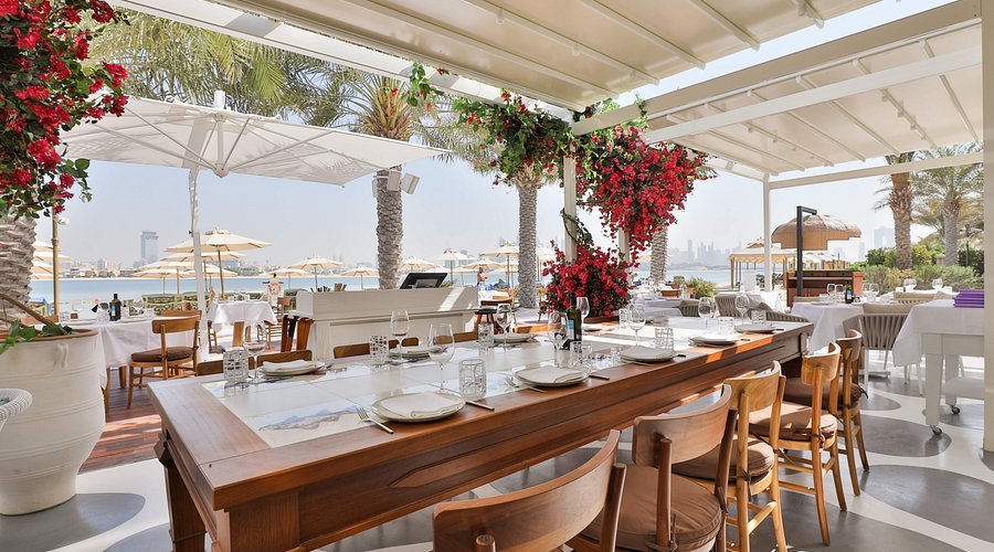 Open-air waterfront restaurant with set tables, wooden chairs, palm trees, and red flowers under a white canopy