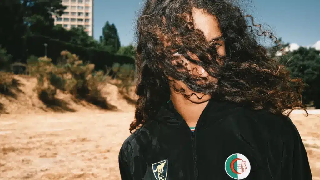 Person in a black jacket with curly hair blowing across their face outdoors on a windy day