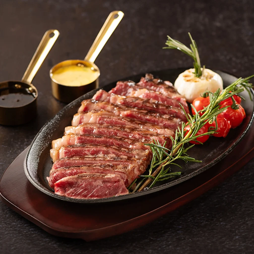 Best Brunch in Dubai: Sliced grilled steak on a cast-iron platter with rosemary, cherry tomatoes, and sauce cups