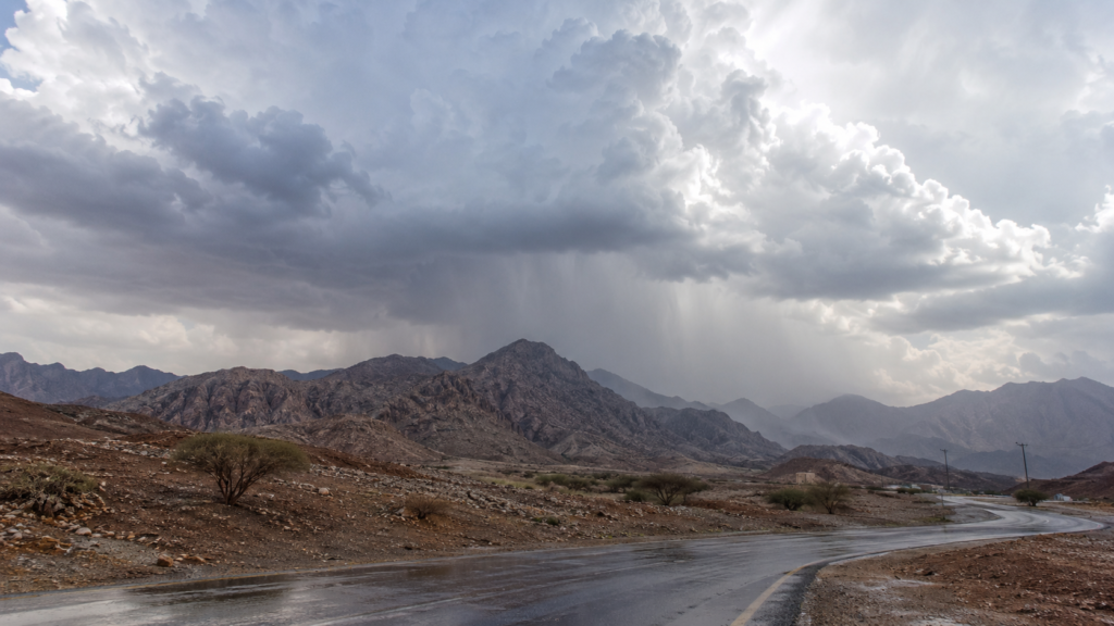 Winding wet road through a rocky desert valley under dramatic rain UAE storm clouds and rain over distant mountains