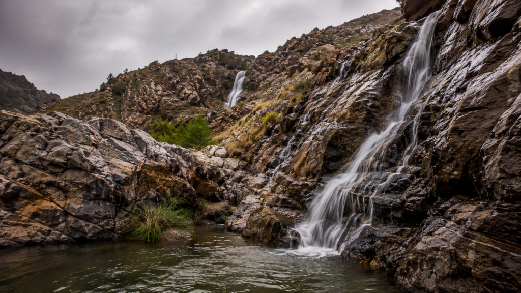 Waterfalls cascading down rocky mountainside into a small pool under a cloudy sky