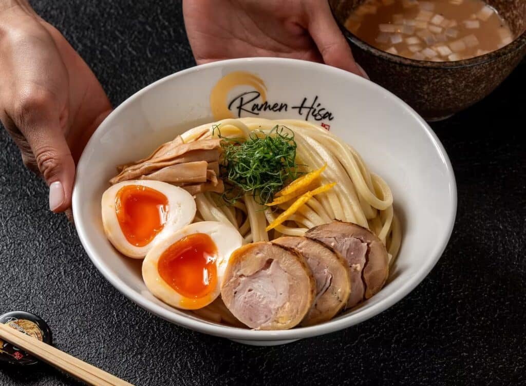 Ramen bowl with soft-boiled eggs, sliced pork, bamboo shoots, herbs, and thick noodles being held over a table