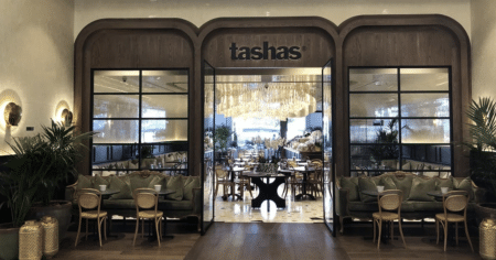 Elegant restaurant entrance with arched wood framing, green sofas, and a large chandelier inside, labeled tashas