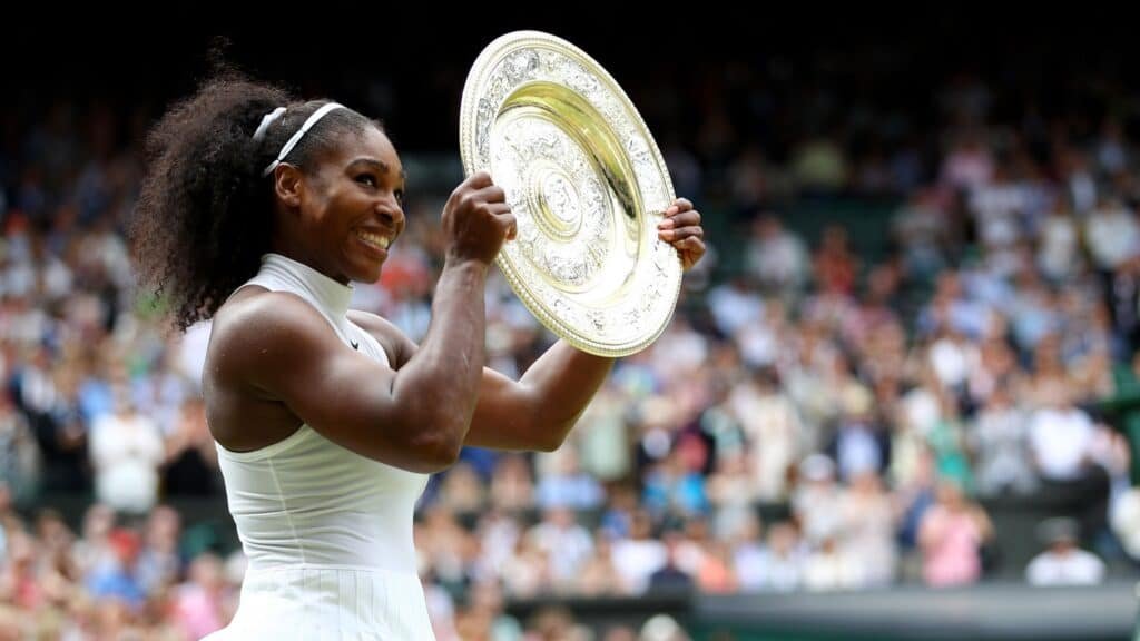 Tennis player Serena Williams smiling and holding up the Wimbledon championship trophy after a win