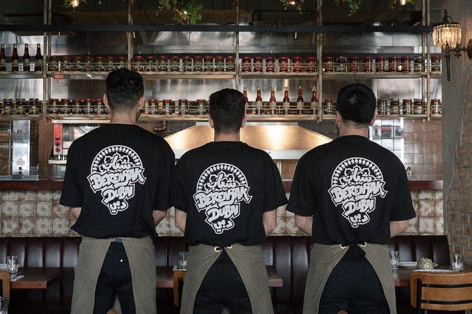 Three restaurant staff members in black logo T-shirts and aprons stand facing a kitchen counter