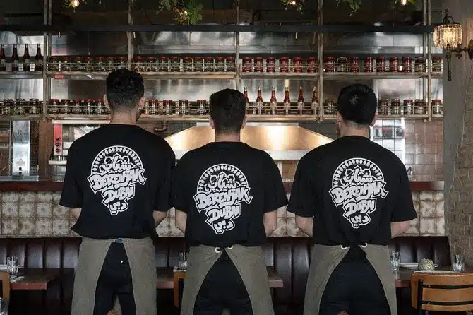 Three restaurant staff members in black logo T-shirts and aprons stand facing a kitchen counter