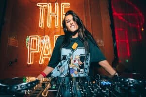 Woman DJ smiling behind a mixing console in a neon-lit club with a sign reading THE PA