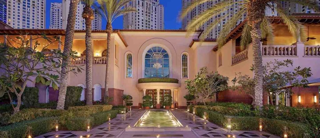 Luxury courtyard at dusk with palm trees, a lit reflecting pool, and an elegant building facade