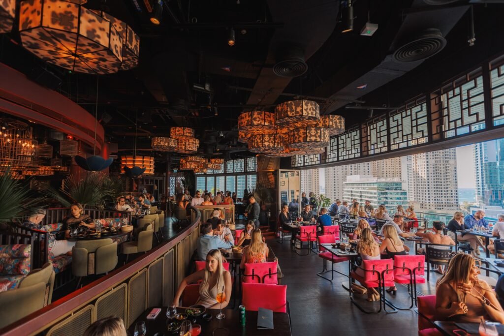 Ladies Night: Busy upscale rooftop restaurant with patterned pendant lights, pink chairs, and city views through large windows