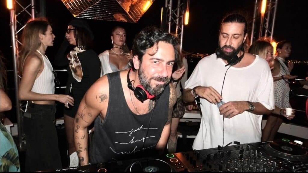 Jixo & Danz at a nighttime party, with people mingling behind them