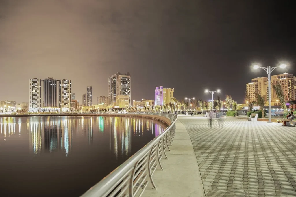 Ajman Corniche with city skyscrapers reflected in calm water and lit streetlamps along the walkway