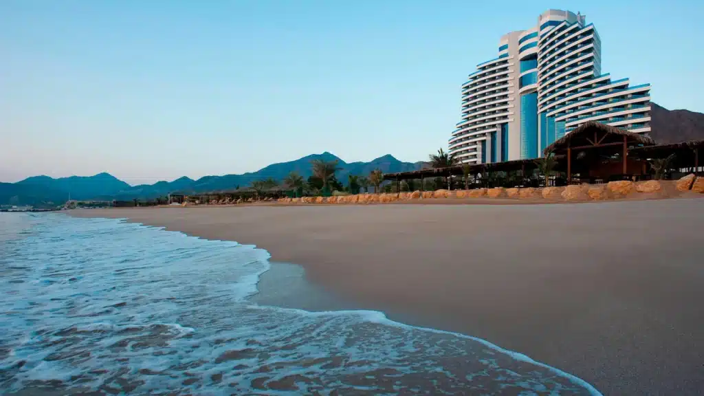 Curved Al Aqah beachfront hotel beside a quiet sandy shore with waves, palm trees, and mountains in the background