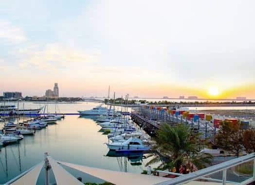 Al Hamra Marina with docked yachts at sunset, calm water, and a colorful pier under a bright sky