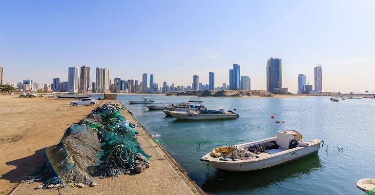 Small fishing boats moored by a harbor beside piled nets, with Al Khan skyline across the water