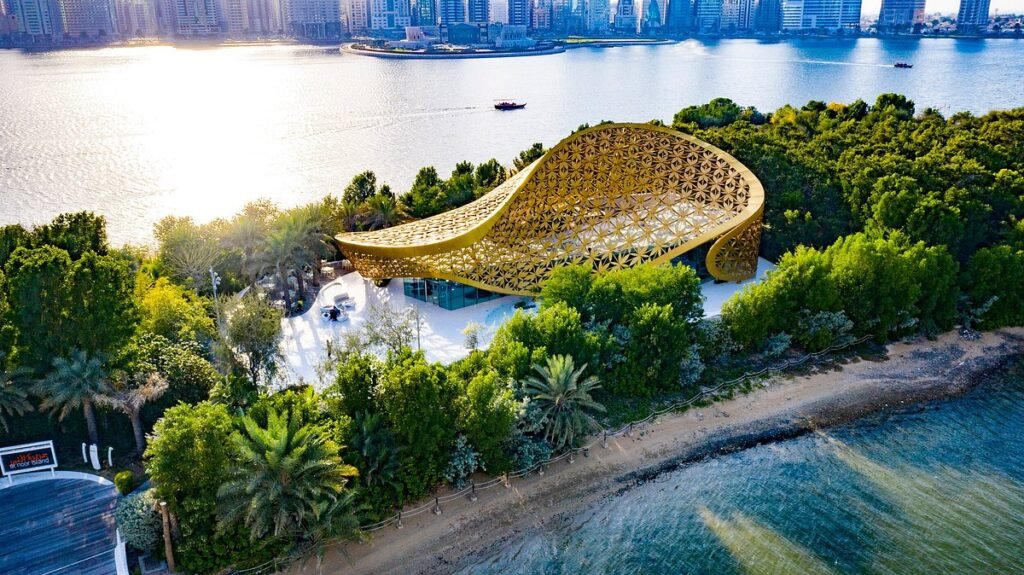 A golden lattice pavilion sits among lush trees on a waterfront with a city skyline in the background